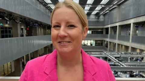 Hannah Bardell wearing a bright pink blazer stands in the foreground of a large modern atrium with multiple floors, metal railings, and overhead skylights. The spacious interior features grey architectural panels, exposed walkways, and conference or office spaces visible in the background.