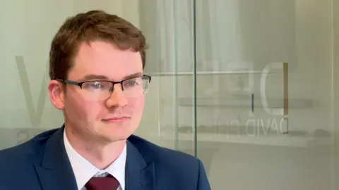 Lawyer Aiden Walley is starring off camera at the interviewer. He is wearing glasses and a blue suit with a white shirt and burgundy tie.