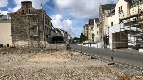 The former CI Tyres site is level and gravelled. There is a metal fence around it. There are houses next to and opposite it.