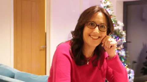 Christina Reeves sits at a table with one of her hands resting on her face. She is smiling. She has long wavy brown hair and wears a pink jumper. A Christmas tree with lights on can be seen behind her and it is reflected in some French doors.
