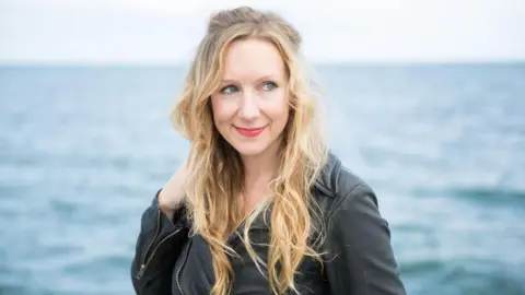 Katie Dutton A woman with blonde hair, wearing a black jacket, stood in front of the sea