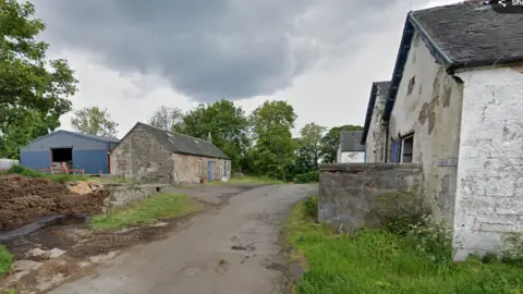 A road leading into a farm, with several stone farm buildings visible. Grass and mud are around the road.
