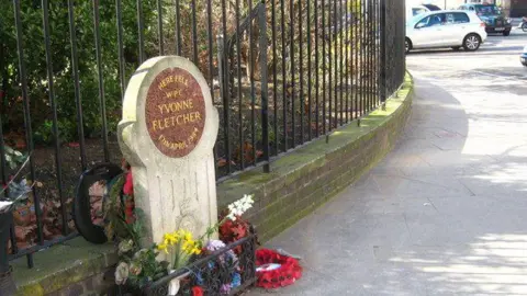 Rod Allday A white stone memorial with a curved top and floral tributes at its base carries the words "Here fell WPC Yvonne Fletcher 17th April 1984"