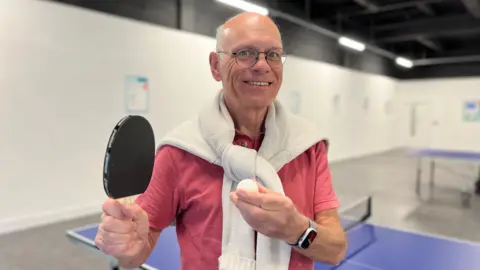 Head and shoulders image of Tim Bick, holding a ping pong bat in his right and ball in his left. Bick has rounded glasses and is wearing a white jumper over his shoulders and a pink shirt.