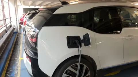 A white hatchback is reverse parked in an underground car park. It is being charged by an electric vehicle charger, which is connected to its charging point.