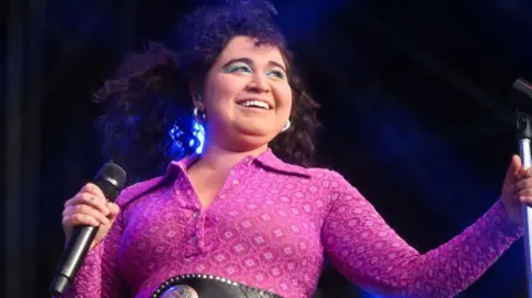 Getty Images Remi Wolf on stage. Remi has curly brown hair and bright blue eye makeup. She wears a patterned pink jumpsuit and smiles at the crowd. 