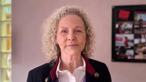 Dr Jacqui Montgomery-Devlin. She is an older woman with curly, blonde hair. She is looking at the camera with a neutral expression on her face. She is stood in a room with a pale pink wall, and a framed photo. She is wearing a white shirt, with a black blazer and red collar.
