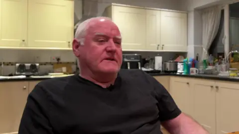 A man wearing a black t-shirt sits at a kitchen table. A kitchen surface top, cupboards and utensils can be seen behind him.