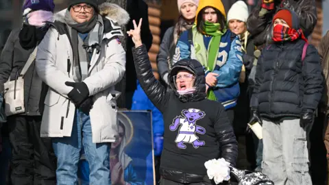 A woman wearing a black puffer jacket with a purple bear motif puts her right arm in the air in a peace sign, at a makeshift memorial for Alex Pretti in south Minneapolis.