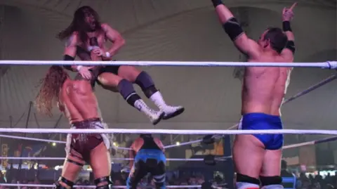 Johnny Jupiter Four wrestlers inside a ring. One with his back to us is lifting another in the air around the throat and is about to slam him down. Another man is on the right with both hands raised in the air. A fourth man is slumped over the ropes on the far side. 