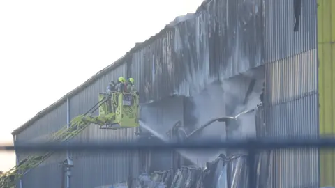 PA Media Two firefighters in high-visibility protective gear stand in the basket of a turntable ladder, directing a water jet into a large hole in the charred, corrugated metal wall of an industrial building. Smoke billows from the damaged structure.