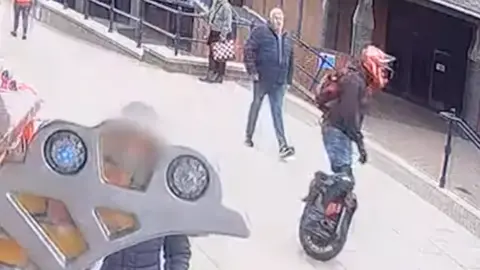 A man in a hooded black top or coat, a red helmet and blue jeans riding on an electric unicycle on a pedestrianised area.