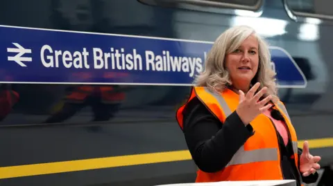 PA Media Heidi Alexander in an orange high-vis jacket in front of a train which has 'Great British Railways' written on it.