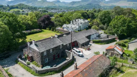 Lake District Calvert Trust An aerial view of a grey-brick building with a house on one side, which extends into a barn. The doors are painted red and it is surrounded by two stone walls. There are a few similar buildings, some are red and some are painted white, around it. Beyond there are the fields and trees of the countryside, with fells in the distance.