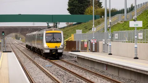 Network Rail A yellow and grey train made up of three carriages is pulling into an empty platform two at a station on the Marston Vale line. The driver is wearing orange hi-vis and there is no destination listed at the front of the train.