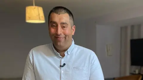 Tauseef stands and smiles in a living room with a lamp light see on behind him. He is wearing a blue shirt with a black lapel mic attached.