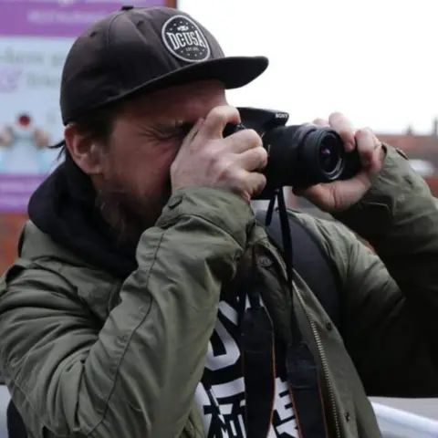 A photographer looks through the viewfinder of a camera while holding it up to his face with both hands. His right eye is closed. He wears a black baseball cap and a green coat over a black and white printed shirt.
