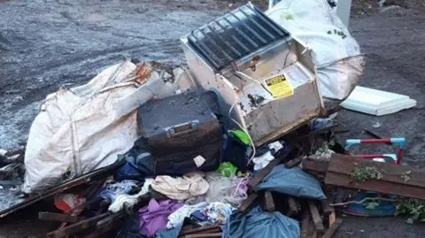 North Northamptonshire Council A large collection of rubbish, showing a fridge, suitcase, items in bags, sacks and pieces of wood, all on the ground. 