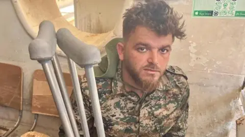 A young man dressed in a camouflage combat uniform holding crutches stares at the camera. His face is dirty and his eyes look tired.