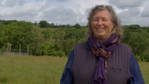 Dorette Engi, smiling and wearing a blue jumper with purple scarf and gillet. She is standing in her land with the stork avery in the background.