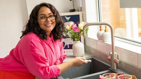 Shelina Permalloo leaning over a sink in her kitchen and smiling. She is wearing a pink top with a red apron.