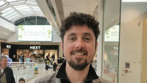 A man stands in front of several shops in a shopping centre. He is smiling, wearing a grey jacket and has short dark hair and a beard.