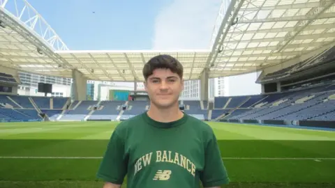 Meilir Rowlands A young man with brown hair, wearing a green t shirt with the words NEW BALANCE written across the front in beige writing. He is smiling and looking at the camera and is stood with a football pitch and stands behind him.