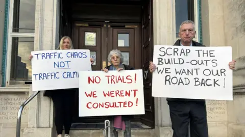 LDRS Two women and a man stand in front of a building and hold signs towards the camera. One says: "Traffic chaos. Not traffic calming." The middle sign reads: "No trials! We weren't consulted!" The sign on the right says: "Scrap the build-outs. We want our road back!"