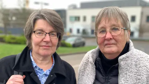 Pictured are Neighbours Pamela Wood with short dark hair, black glasses and is wearing a flowery blue shirt, she is standing new to Sue Bowdery who has short light hair and is wearing think framed glasses, a black zip-up jacket and grey fluffy coat.