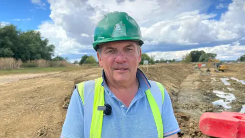 Andy Timbrell standing in front of a trench which has been dug to make way for the new canal. He is wearing a hard hat and a high-vis vest over a blue polo shirt, and smiles at the camera.