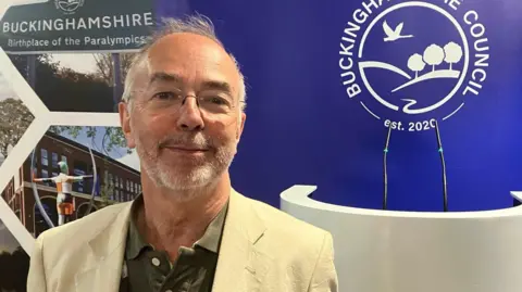 Martin Tett a man who is looking at the camera and smiling and is wearing a cream suit jacket and khaki shirt. He is standing in front of a blue board which says BUCKINGHAMSHIRE COUNCIL in white writing.