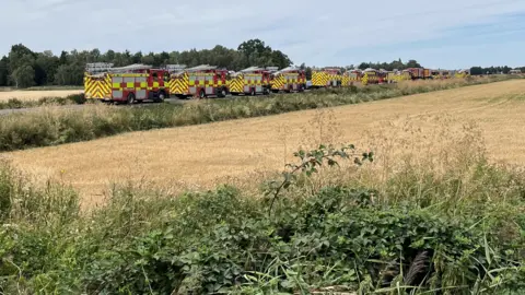 John Devine/BBC A long line of red fire engines parked in a line on a road that crosses fields. Trees can be seen in the distance.