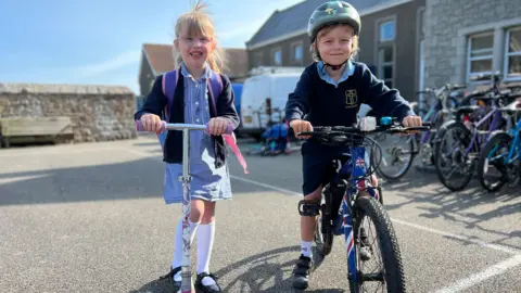 Two children outdoors in a school playground. One with a bike the other with a scooter. Both are dressed in school uniform, with the one on the bike wearing a green helmet.