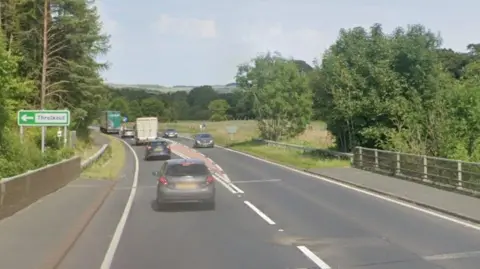 Threlkeld Bridge. It is a two-lane bridge with pavements and grey parapets on both sides. Three cars and two lorries are travelling in the left lane. Two cars are travelling in the right lane. There is a green sign reading Threlkeld and pointing left.