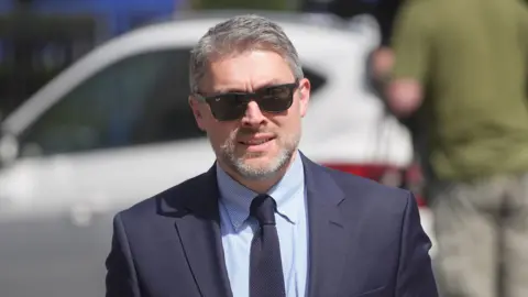 PA Media Adam Smyth, Director for BBC Northern Ireland, wears black Ray Ban sunglasses. he has short greying hair and a beard. He wears a navy suit, blue shirt and navy tie. 