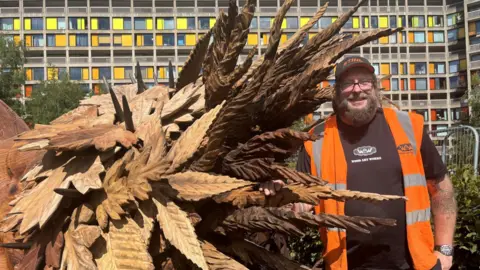 Simon Thake A beaded man in a baseball cap and orange jacket stands next to the head of a wooden sculpture.