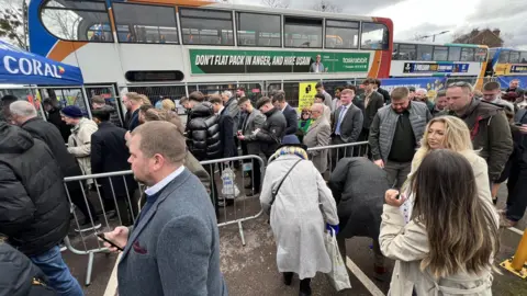 Queues of people attending the Cheltenham Festival are queuing to get on a bus. They are separated by barriers.