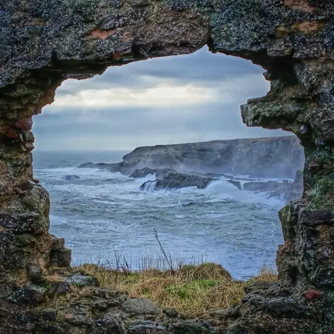 Iain Kelman Waves crashing against the coast, viewed through an old grass-covered stone window arch.