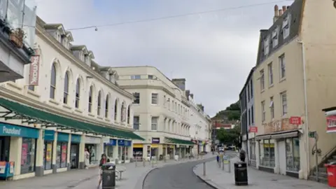 A street scene with shops and a narrow roadway and an awning over one shop and some people walking on the pavement.