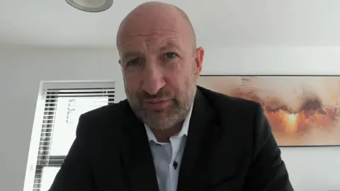 A bald man with a short grey beard, Wearing a white shirt and black blazer, pictured against a white wall with an orange picture in the background.