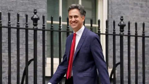 PA Media Ed Miliband in a suit and red tie walking beside a fence 