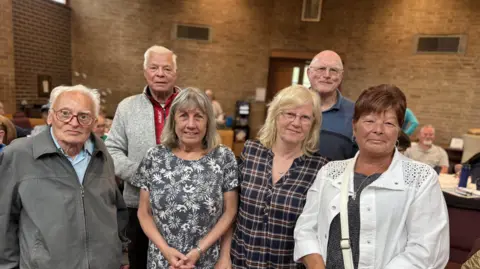 Six South Shields residents posing to the camera. Tom Atkinson is second from left wearing a grey cardigan. He has grey hair. 