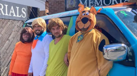 The friends are dressed in their characters and stand by their van or Mystery Machine. The van is blue.