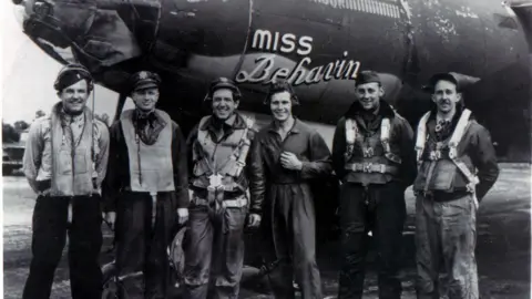 A black and white image of six men standing in front of an aircraft named "Miss Behavin". They are smiling.