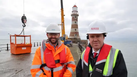Benjamin Leng (left) and Councillor Kevin Johnston standing in front of the lighthouse on Roker Pier. Mr Leng is wearing an orange hi-vis outfit and has a dark beard. Mr Johnston is wearing a black jacket. Both men are also wearing white hard hats.