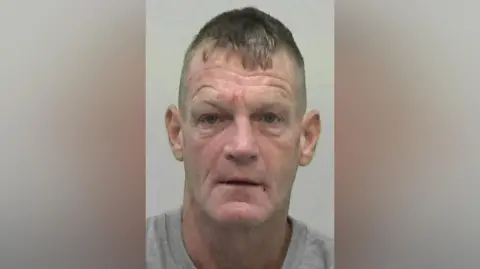 Mugshot of Stephen Thompson. He is clean shaven with short fair hair and deep wrinkles. He has a few marks on his face that look like small but recent wounds.