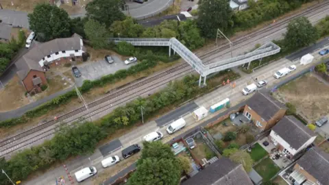 An aerial photo of the railway track in Wickford where a train derailed on Friday. The train has been removed but there are lots of vehicles parked on an adjacent road. There is a footbridge over the track