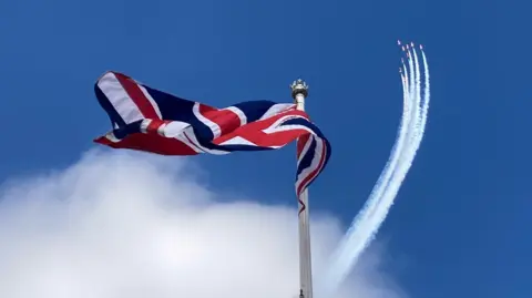 Jake Zuckerman/BBC A union jack flag flying in the sky which is held on a silver pole. You can see the Red Arrows jets to the right in the distance making a formation in the air with a trail of cloud behind them.
