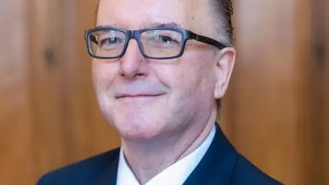 Worcester City Council City council managing director David Blake is seen in an official photograph. He is wearing a white shift and navy blue suit and he is wearing glasses and smilling.