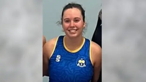 Hannah Cliftlands Hannah Cliftlands, a young women with brown hair in a ponytail wearing a blue and yellow netball kit smiling at the camera. 